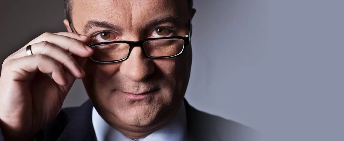 Zbliżenie mężczyzny w okularach, który jedną ręką poprawia szkła i patrzy w obiektyw na jasnoszarym tle.