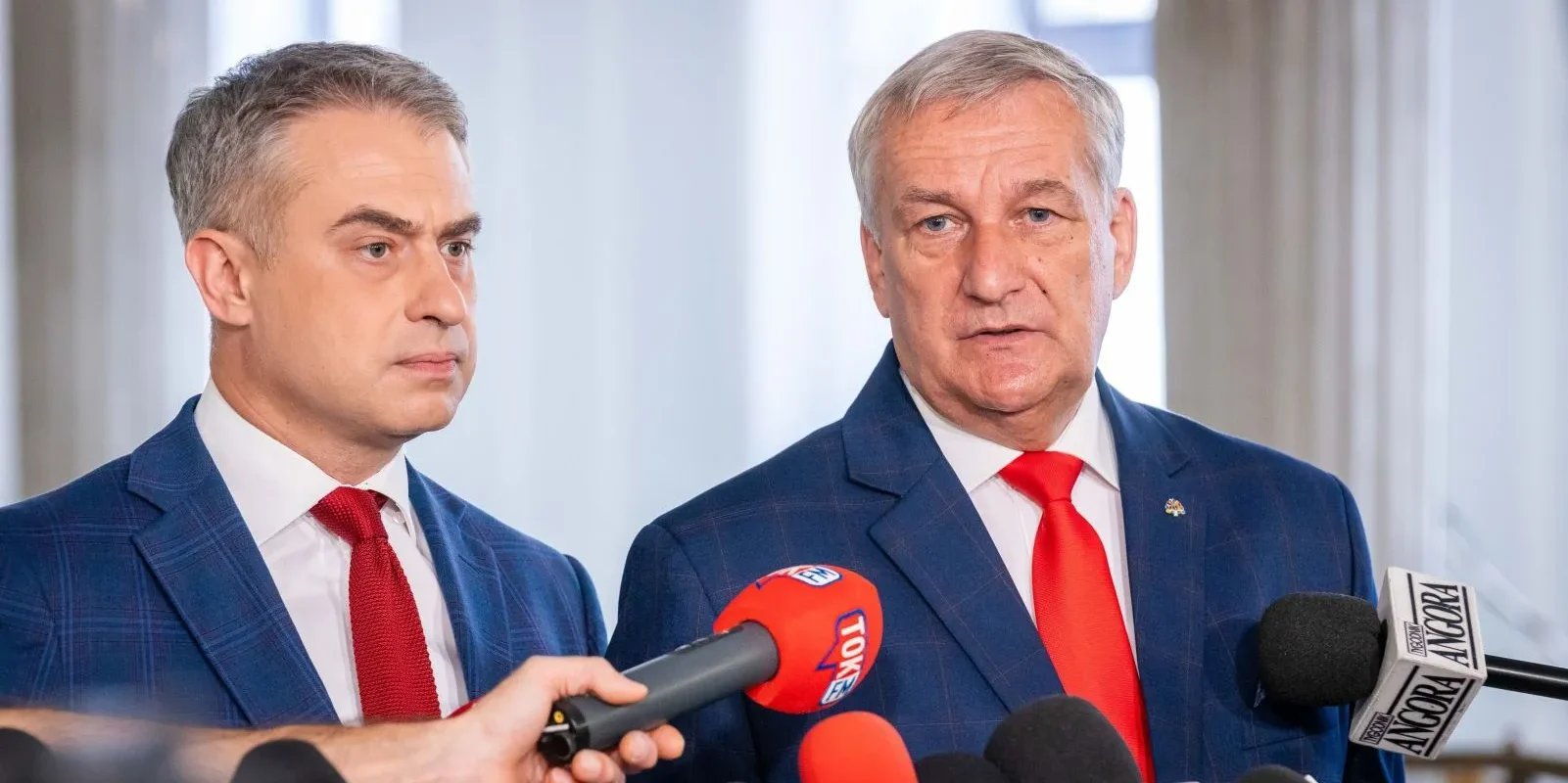 Dwóch mężczyzn w niebieskich garniturach z czerwonymi krawatami udziela konferencji prasowej, stojąc za licznymi mikrofonami.