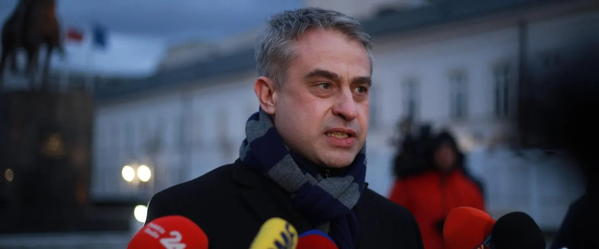 Wieczorem mężczyzna w czarnym płaszczu i szaliku udziela wypowiedzi mediom, stojąc przed mikrofonami, w tle widać posąg konny i budynki.