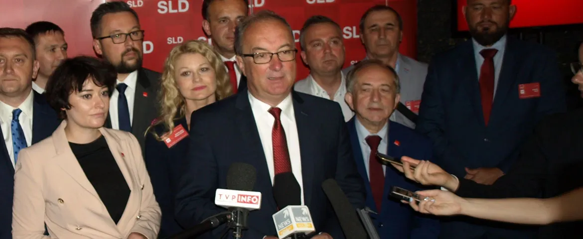 Grupa polityków SLD Lewica Razem na konferencji prasowej; w centrum stoi mężczyzna w garniturze i czerwonym krawacie, mówiący do mediów, otoczony reporterami i kamerami.