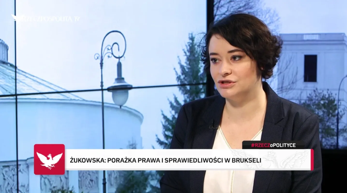 Kobieta w ciemnym żakiecie rozmawia w studiu telewizyjnym, a w tle widać budynek, na dole ekranu widnieje pasek z informacjami.