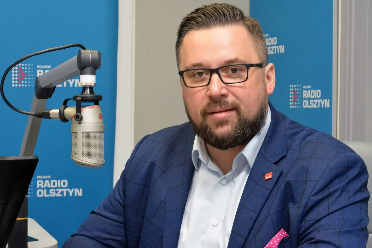 Mężczyzna w granatowym garniturze i okularach siedzi przy mikrofonie w studiu radiowym, na tle niebieskich banerów z logo Polskiego Radia Olsztyn.