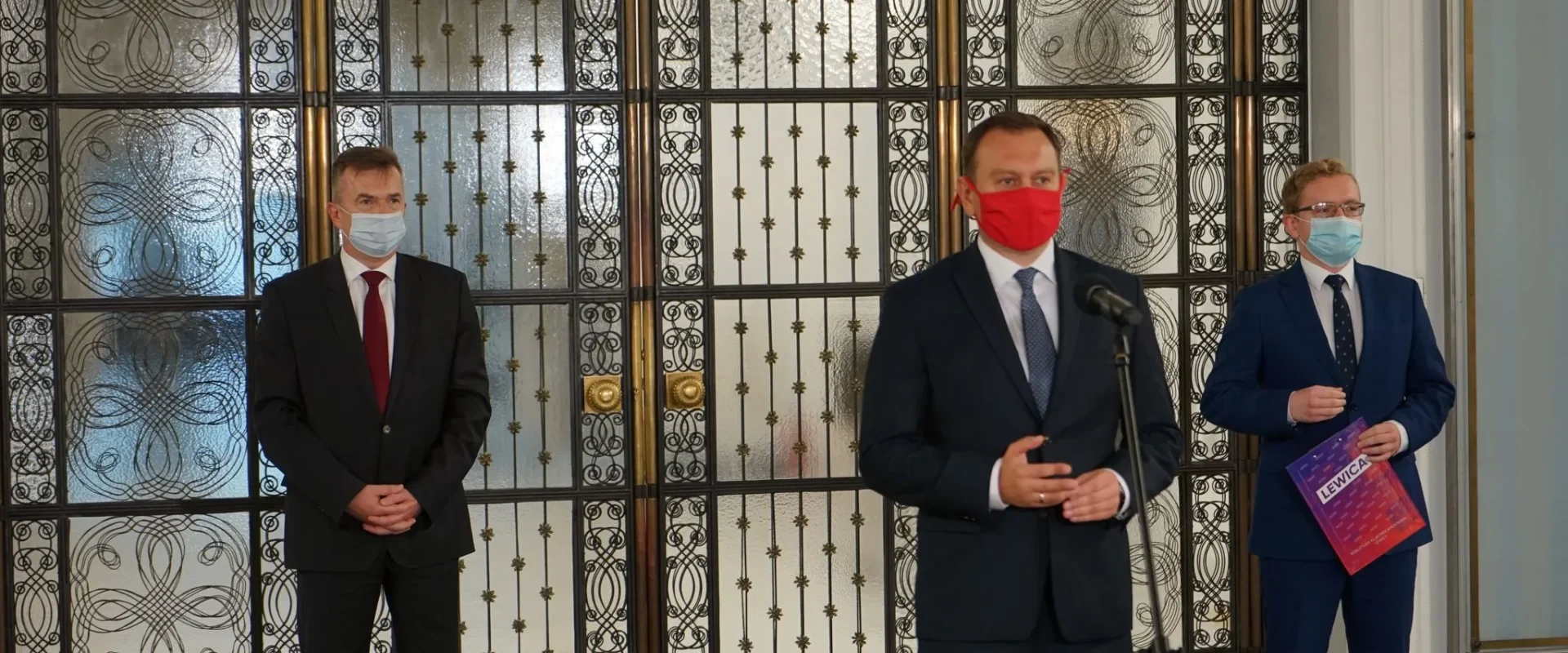 Trzech mężczyzn w garniturach i maseczkach stoi na konferencji prasowej w eleganckiej sali przy zdobionych metalowych drzwiach; środkowy przemawia do mikrofonu, a prawy trzyma broszurę Lewica.