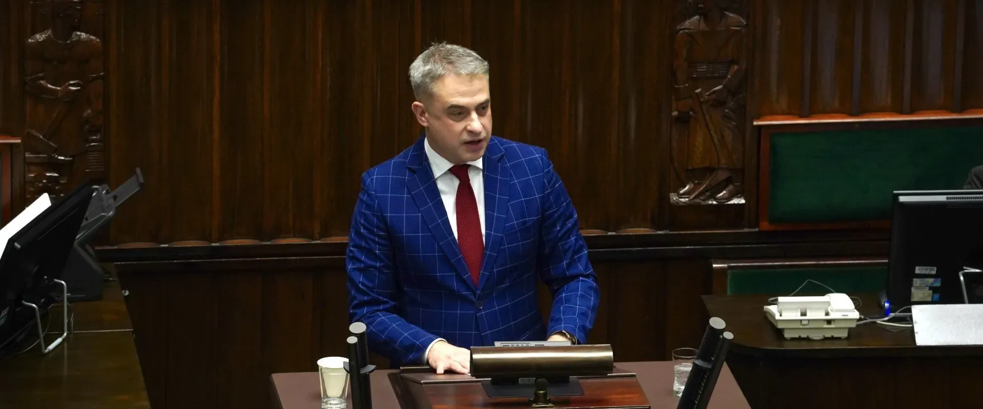 Minister cyfryzacji w niebieskim garniturze w kratę przemawia zza mównicy w sali plenarnej.