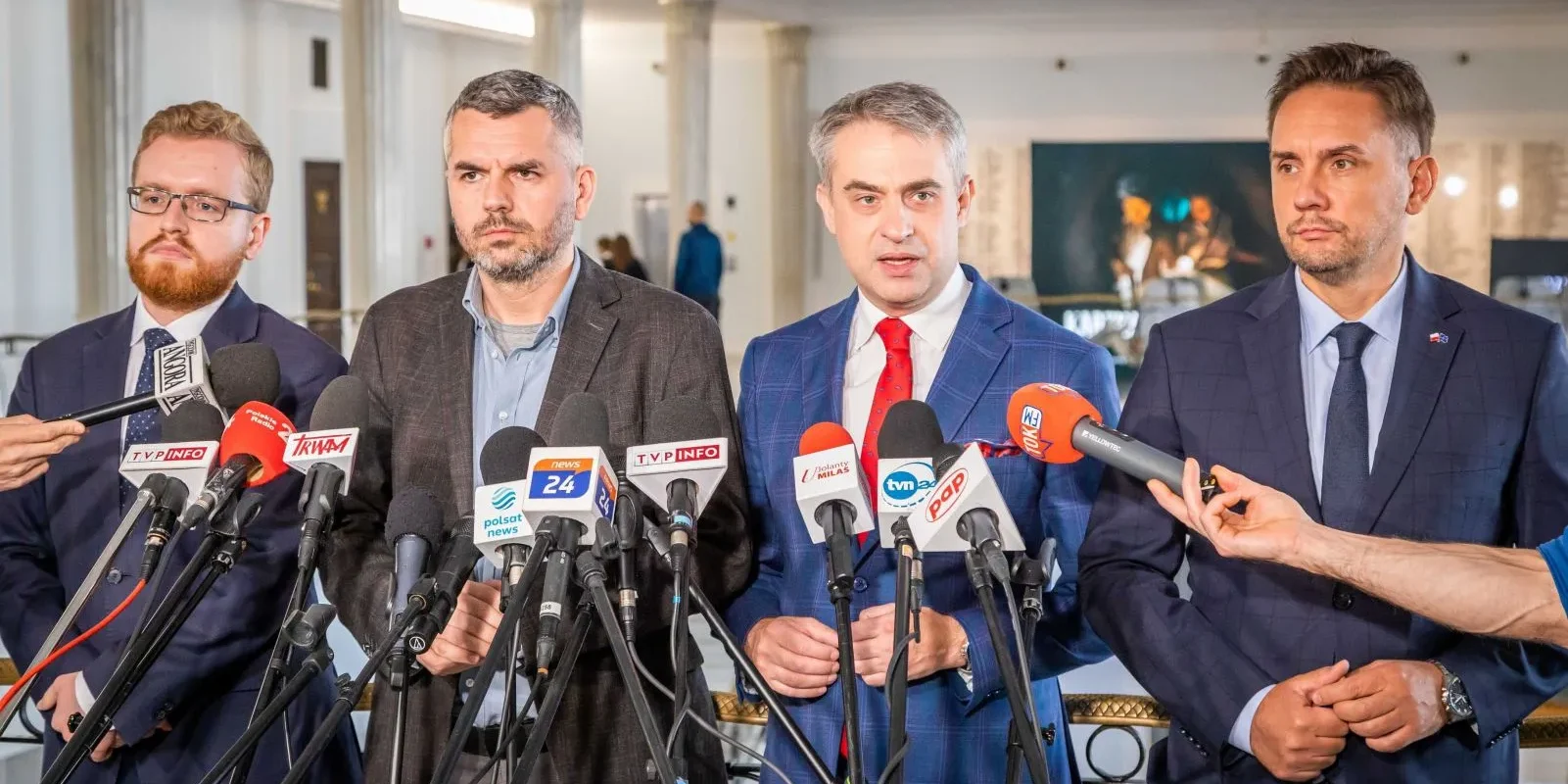 Na zdjęciu czterech mężczyzn w garniturach stoi za mikrofonami i udziela wywiadu w sali przypominającej przestrzeń konferencyjną.