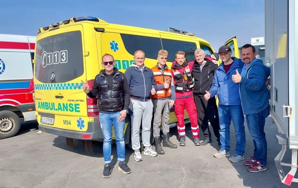 Na zdjęciu widoczni są mężczyźni pozujący przy ambulansie, na tle niebieskiego nieba, uśmiechający się i pokazujący znaki zwycięstwa oraz kciuki w górę.