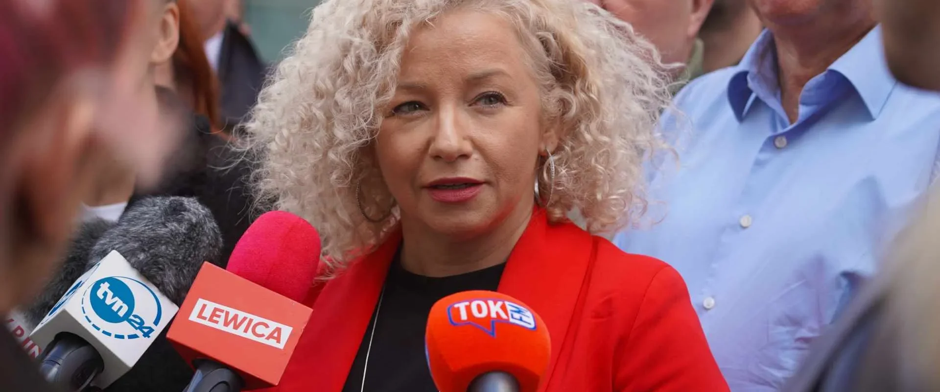 Szefowa sztabu wyborczego o kręconych blond włosach w czerwonej marynarce udziela wywiadu mediom podczas wyborów do Parlamentu Europejskiego, otoczona reporterami i kolorowymi mikrofonami.