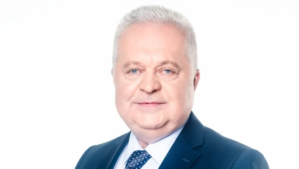 Portret mężczyzny w granatowym garniturze, jasnoniebieskiej koszuli i wzorzystym krawacie na białym tle.