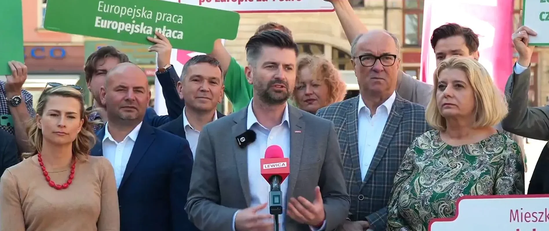 Grupa osób podczas konferencji prasowej Lewicy, z mówcą przy mikrofonie i plakatami promującymi wakacyjne bezpłatne podróże koleją dla osób do 26. roku życia.
