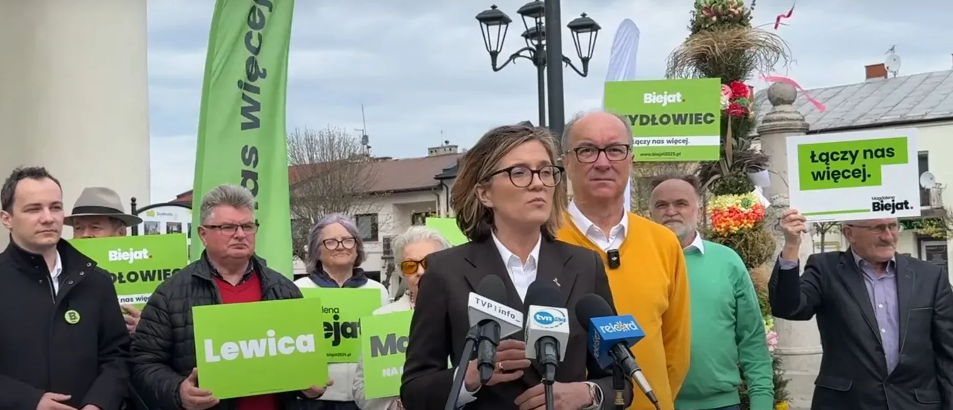 Kobieta w garniturze przemawia na konferencji prasowej Lewicy, otoczona zwolennikami z zielonymi plakatami z napisami Biejat i Lewica, w kontekście planu gospodarczego „100 mld zł na innowacje”.
