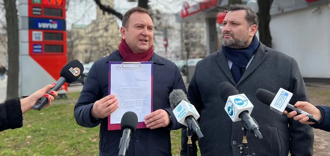 Dwóch mężczyzn w zimowych ubraniach podczas konferencji prasowej na zewnątrz, jeden trzyma klipboard z dokumentem, wokół niego reporterzy z mikrofonami.