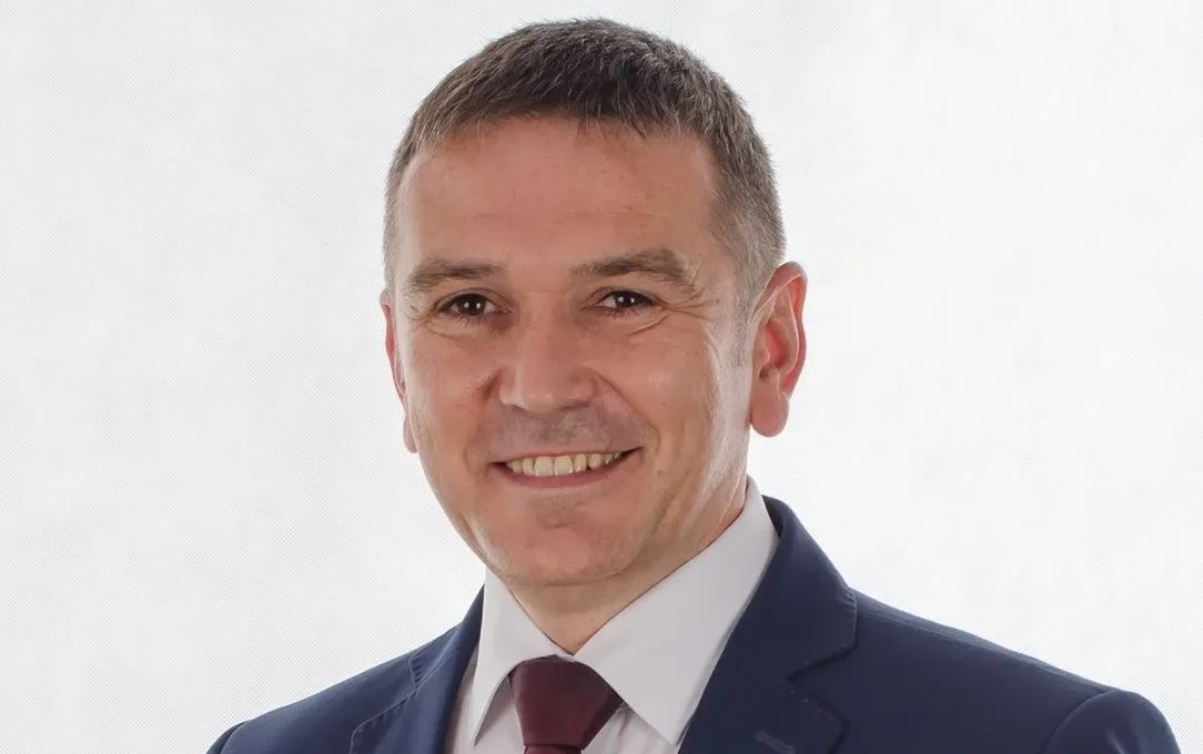 Portret mężczyzny w granatowym garniturze, białej koszuli i czerwonym krawacie, uśmiechniętego na jasnym tle.