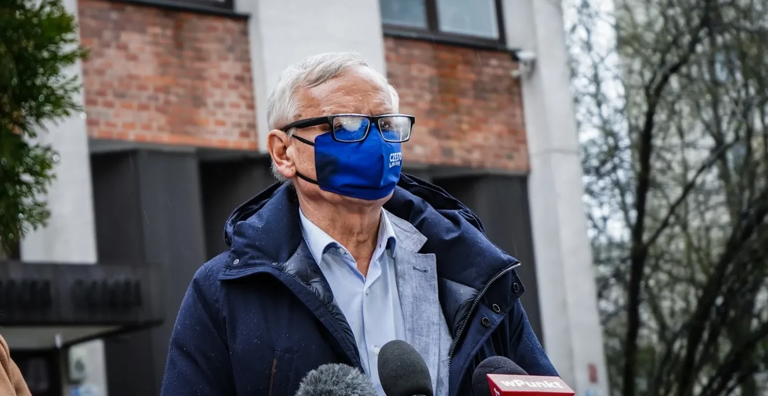 Starszy mężczyzna w okularach i niebieskiej maseczce stoi na zewnątrz przed budynkiem, ubrany w granatową kurtkę i jasnoniebieską koszulę, i przemawia do mediów przy mikrofonach.