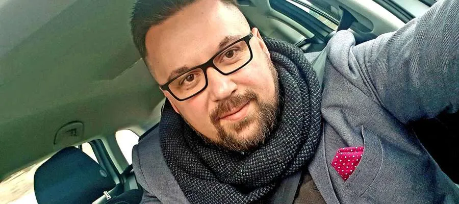 Mężczyzna w okularach i grubym szaliku robiący selfie w samochodzie, ubrany w marynarkę z czerwoną poszetką w białe kropki.