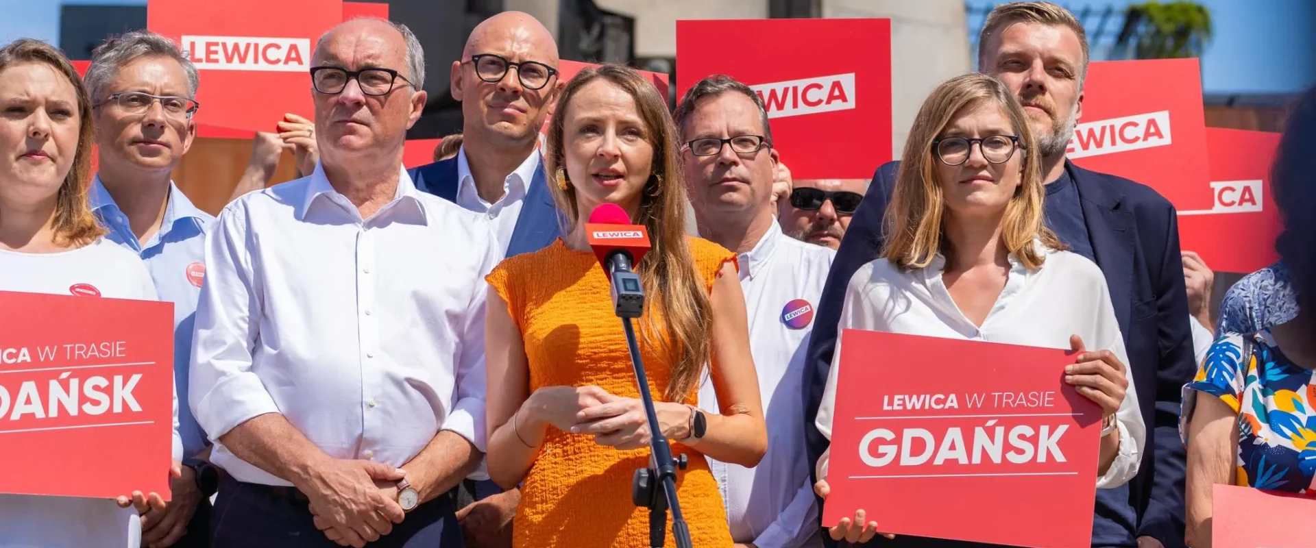 Grupa osób uczestniczy w lewicowej kampanii, trzyma czerwone plakaty z napisem LEWICA, a na środku kobieta w pomarańczowej sukience przemawia do mikrofonu.