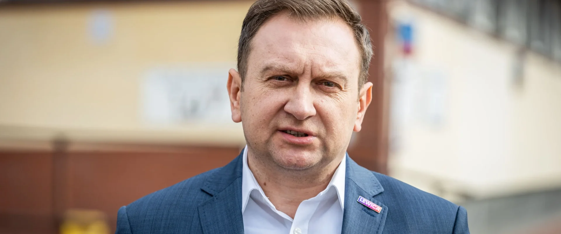 Mężczyzna w granatowym garniturze i białej koszuli stoi na zewnątrz, z przypinką Lewica na klapie, patrząc w kamerę.