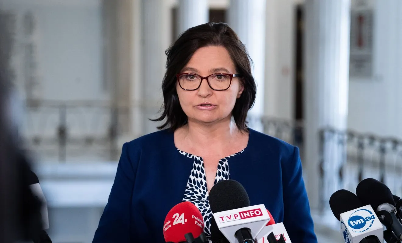 Kobieta w okularach, ubrana w granatową marynarkę, stoi przed mikrofonami podczas konferencji prasowej w jasnym wnętrzu.