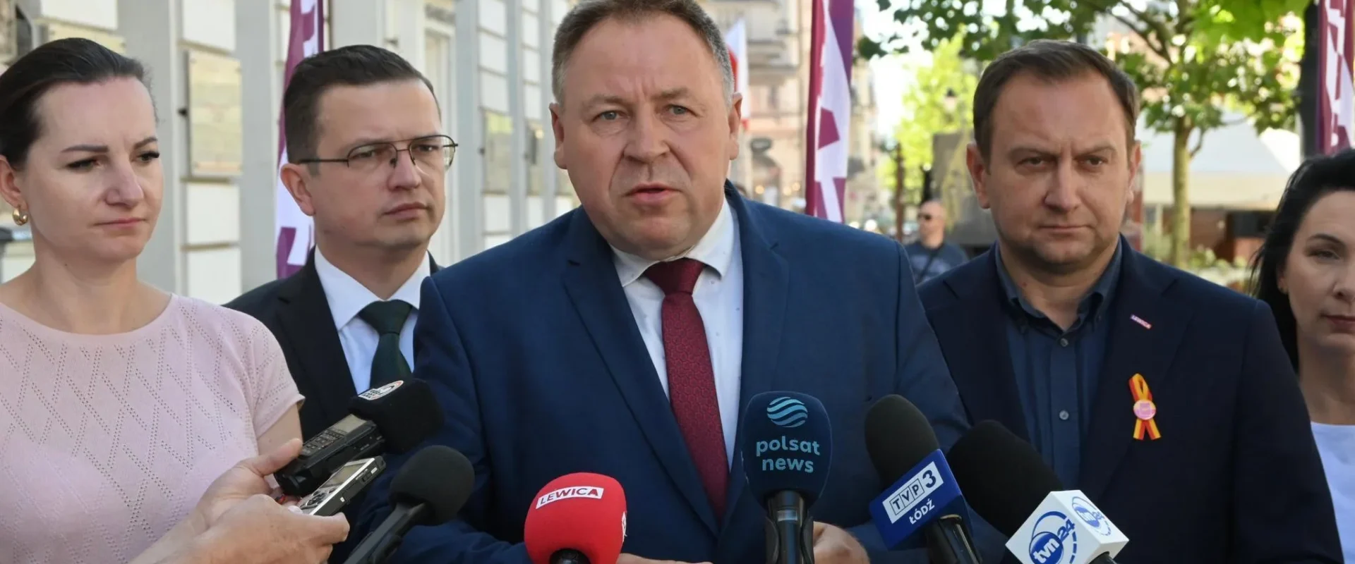 Mężczyzna w granatowym garniturze z czerwonym krawatem wygłasza oświadczenie podczas konferencji prasowej na ulicy, otoczony dziennikarzami i mikrofonami z różnych mediów.