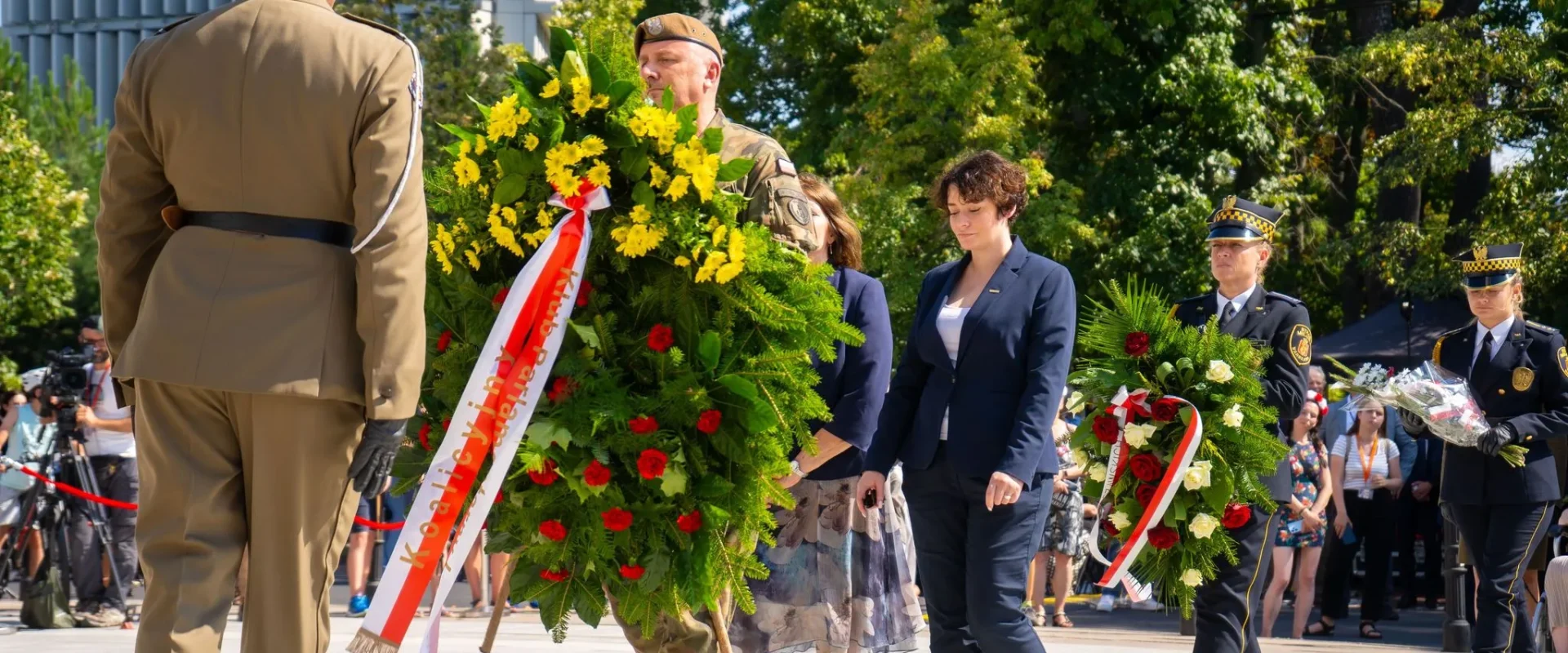Uroczystość upamiętniająca 80. rocznicę Powstania Warszawskiego, podczas której żołnierze w mundurach niosą i składają duże wieńce z żółtych kwiatów przed zgromadzonymi widzami.