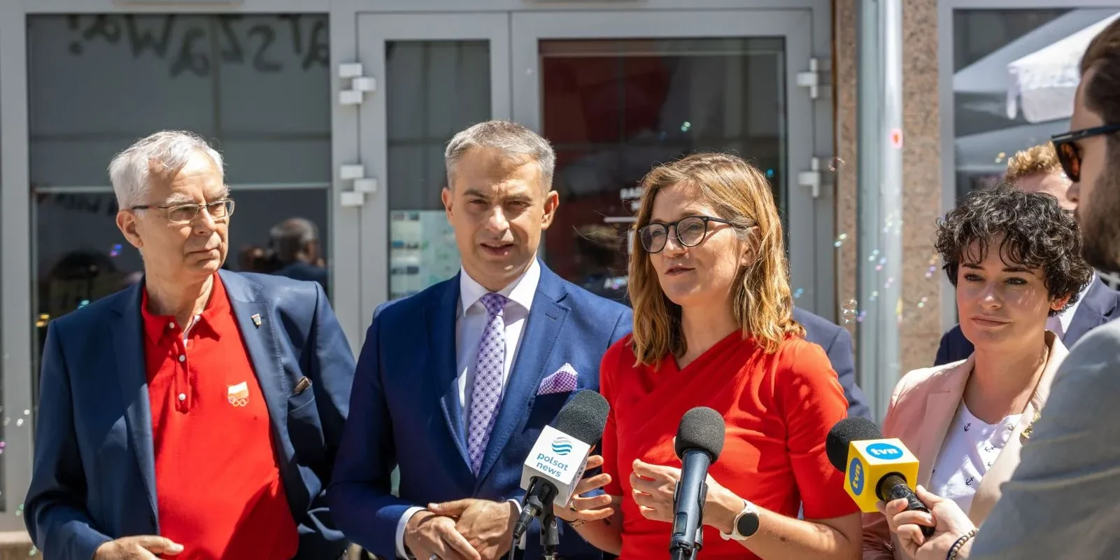 Grupa dorosłych w garniturach i eleganckich ubraniach udziela konferencji prasowej na zewnątrz budynku, stojąc przed mikrofonami z okazji Dnia Dziecka.