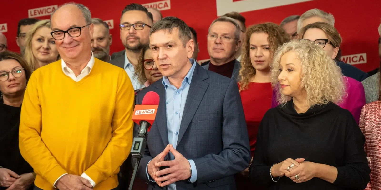 Grupa polityków Lewicy podczas konferencji prasowej; mężczyzna w granatowym garniturze przemawia do mikrofonu z logo LEWICA, otoczony innymi osobami na czerwonym tle.