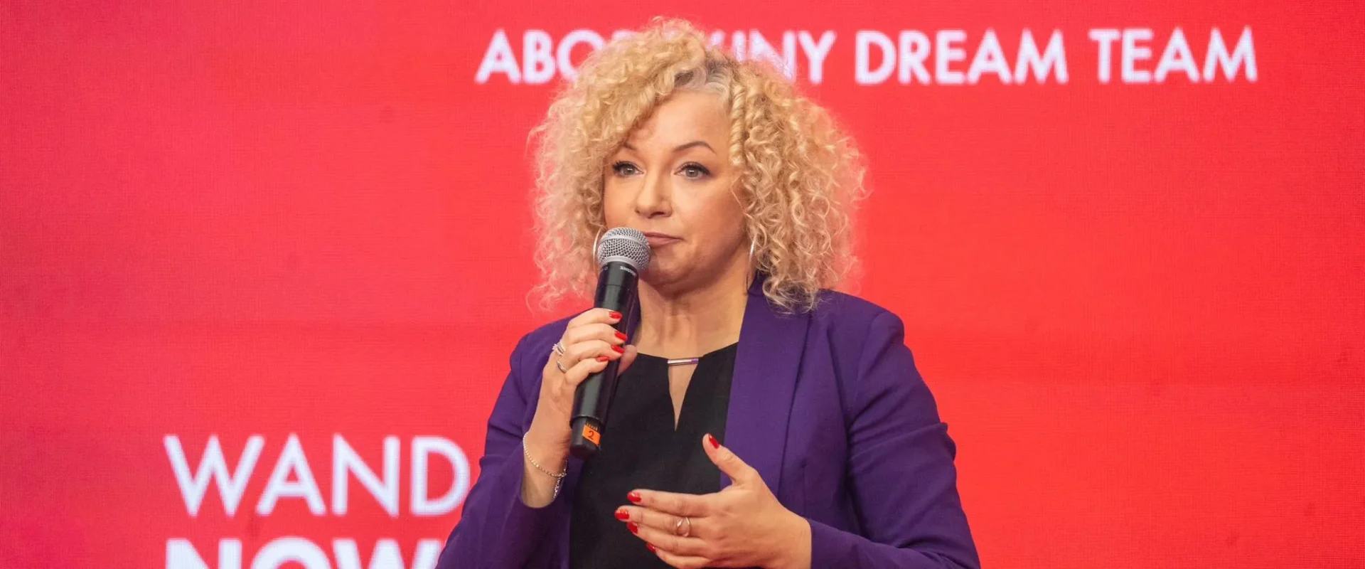 Kobieta o kręconych blond włosach w fioletowym żakiecie przemawia do mikrofonu na czerwonym tle z białymi napisami.
