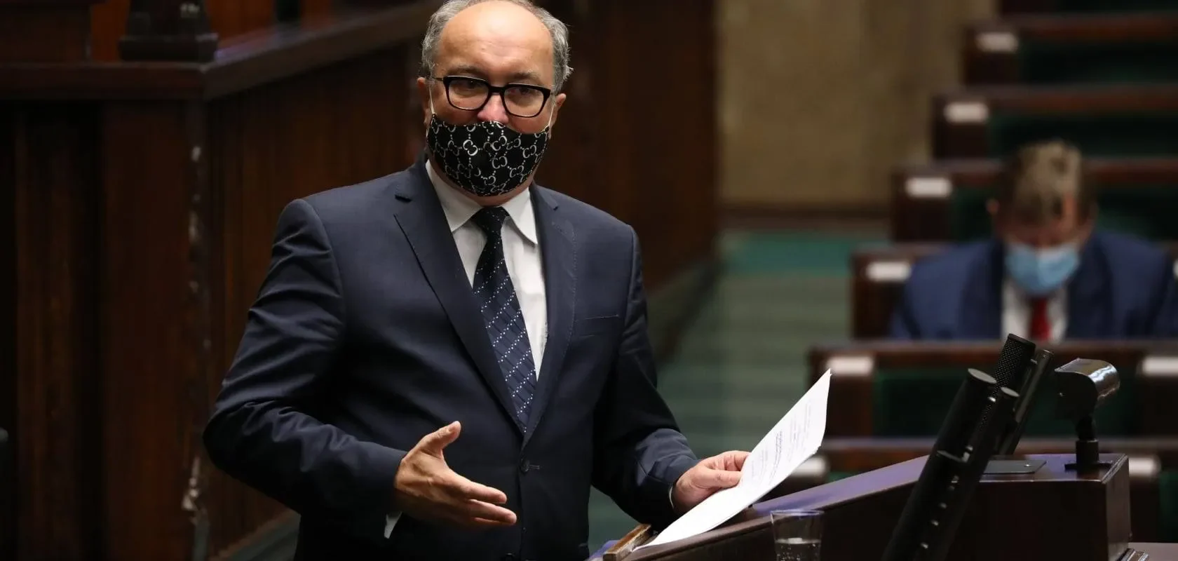 Polityk w garniturze i maseczce z wzorem przemawia z mównicy w sali plenarnej, obok stoją mikrofony.