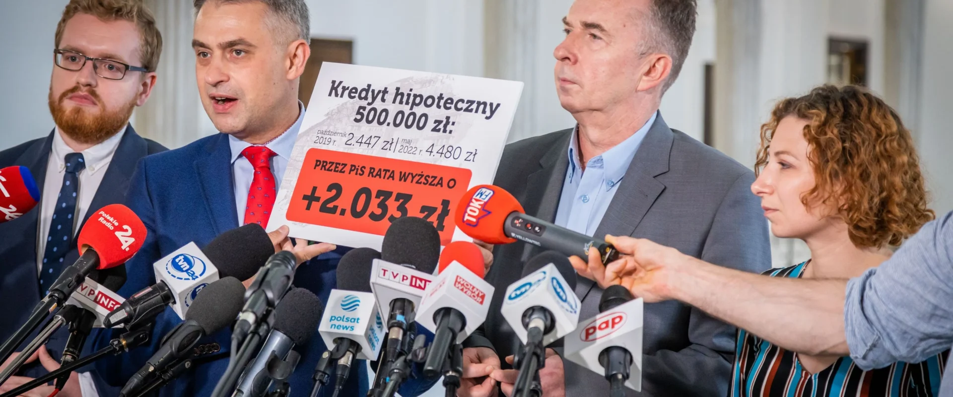 Na zdjęciu znajduje się konferencja prasowa, podczas której przedstawiciele mówią o wzroście raty kredytu hipotecznego, trzymając plakaty z odpowiednimi danymi.
