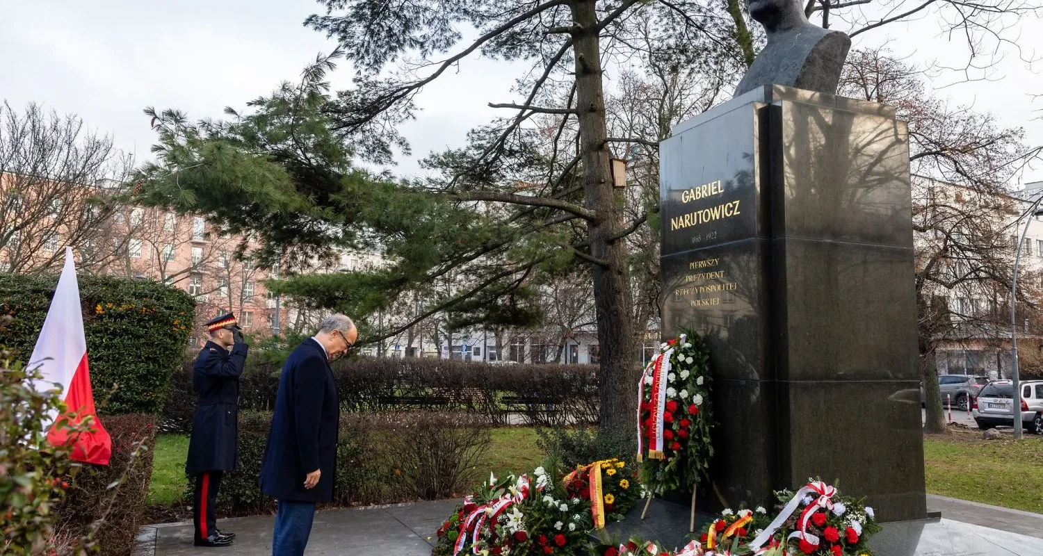 Pomnikowy bust Gabriela Narutowicza w parku z złożonymi wieńcami kwiatowymi oraz dwoma osobami w formalnych strojach oddającymi hołd podczas 103. rocznicy zabójstwa prezydenta.