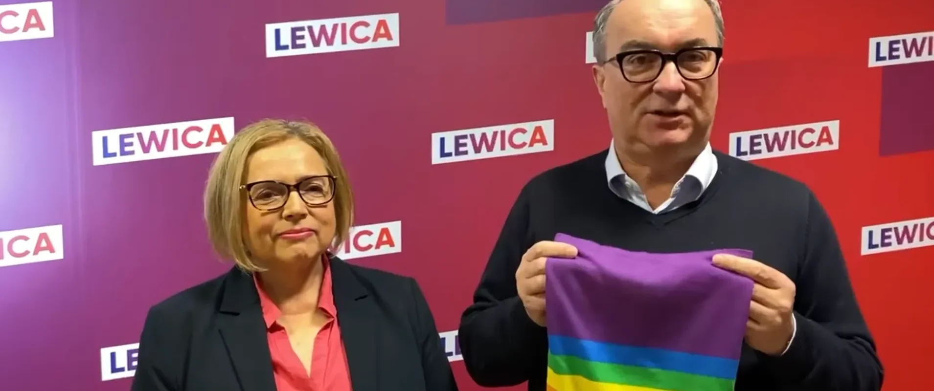 Dwóch ludzi stoi na tle kolorowej ścianki z logotypami Lewicy; mężczyzna trzyma tęczowy komin jako gest wsparcia dla LGBT, wystawiony na aukcję WOŚP.