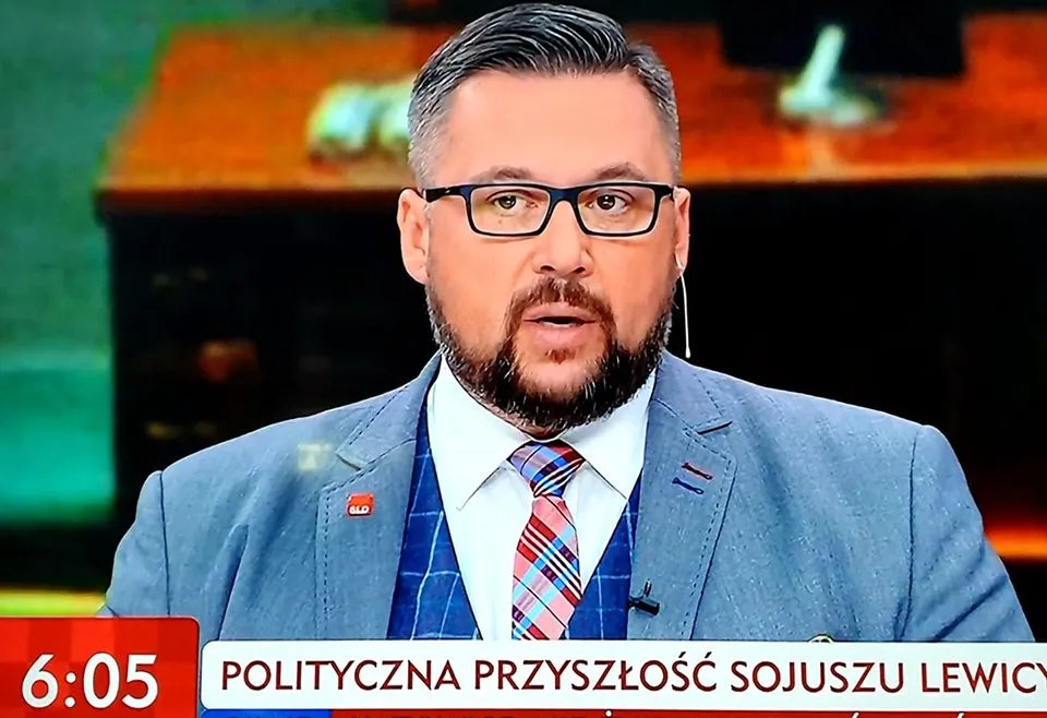 Mężczyzna w jasnoniebieskim garniturze z okularami i krótką brodą przemawia w studiu telewizyjnym, z mikrofonem lavalierowym przy koszuli.