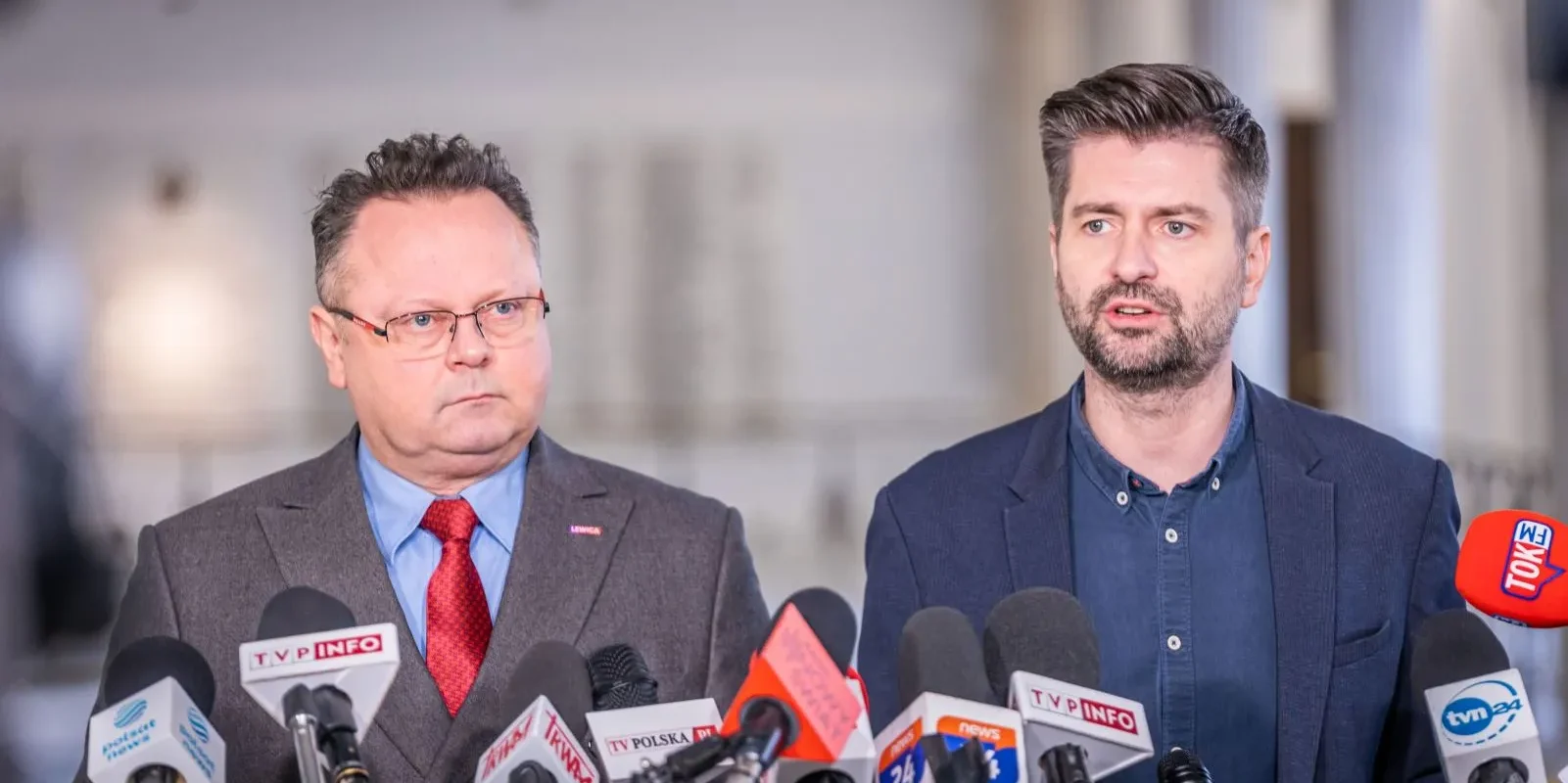 Dwóch mężczyzn w garniturach podczas konferencji prasowej, stojących wśród licznych mikrofonów z logo różnych mediów.