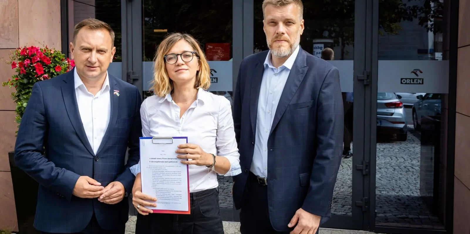 Na zdjęciu widoczni są trzej ludzie stojący przed budynkiem, trzymający dokumenty oraz uśmiechający się do kamery.