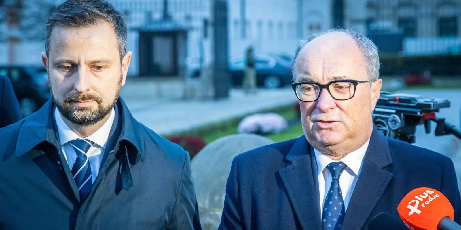 Na zdjęciu widać dwóch mężczyzn w garniturach, stojących na tle budynku, prawdopodobnie podczas wywiadu lub konferencji prasowej.