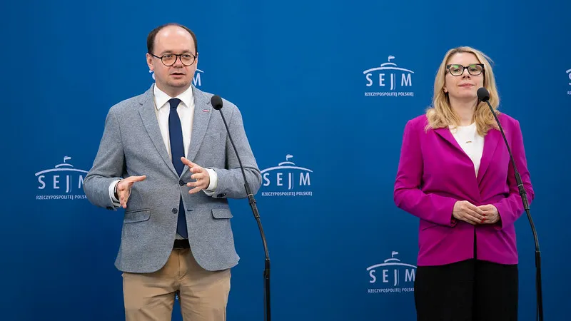 Dwóch mówców stoi przy mikrofonach na niebieskim tle z logo Sejmu; po lewej mężczyzna w szarym żakiecie i beżowych spodniach, po prawej kobieta w różowej marynarce.