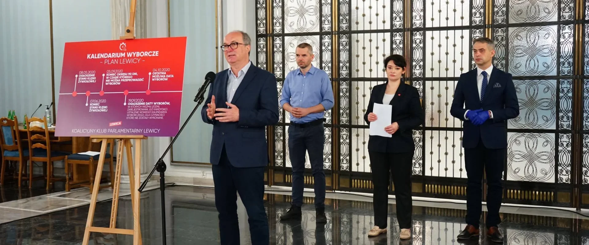 Mężczyzna w garniturze przemawia do mikrofonu przy plakacie wyborczym na sztaludze, a za nim stoją trzy osoby w formalnych ubraniach w eleganckiej sali z dekoracyjną metalową kratą w tle.
