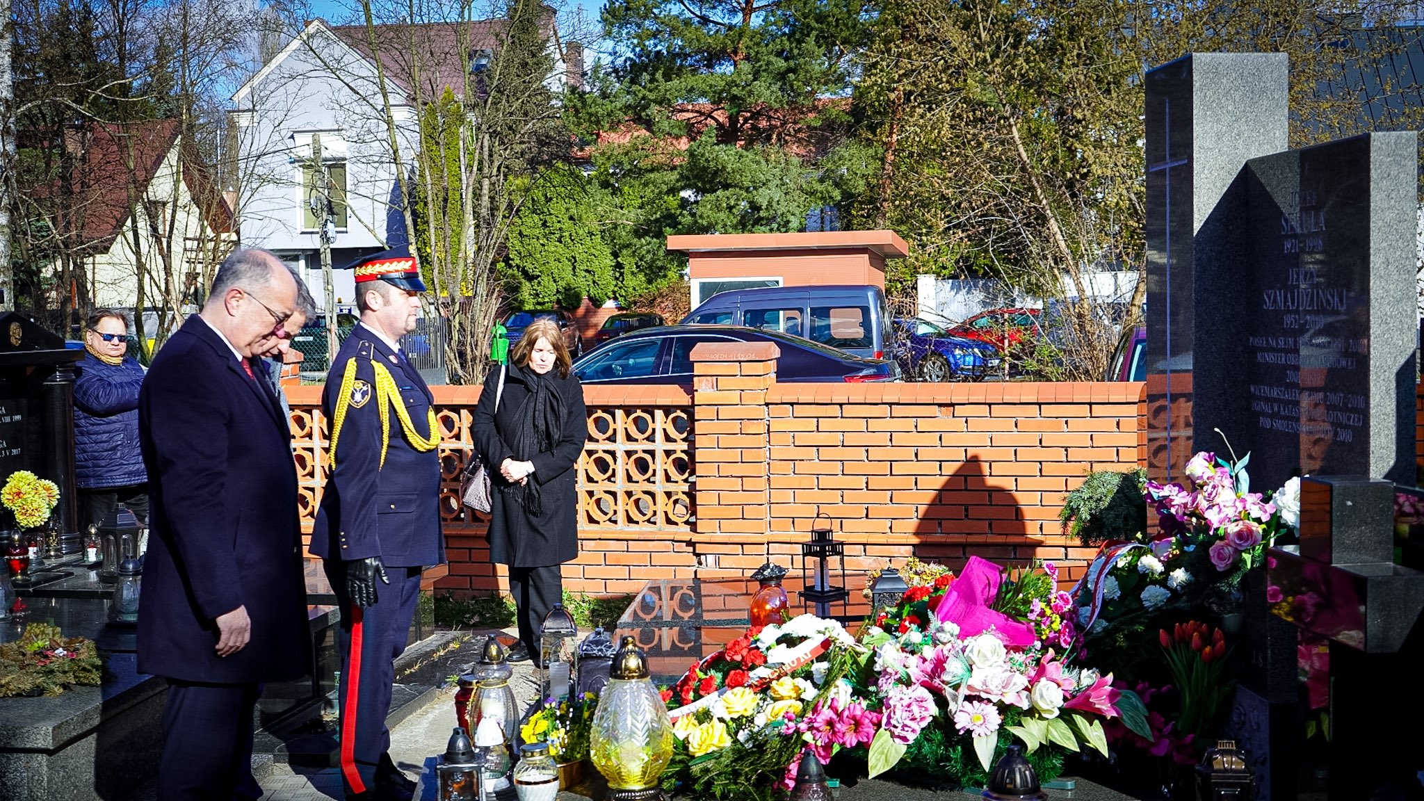 Grupa osób składa kwiaty i modli się przy grobie upamiętniającym tragedię smoleńską; wokół leżą znicze i bukiety, a w tle stoi wysoki granitowy pomnik.