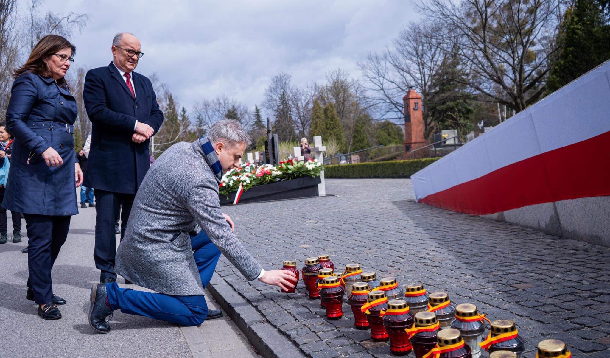 Na zdjęciu widoczna jest grupa osób składających znicze na cmentarzu w celu uczczenia pamięci zmarłych.