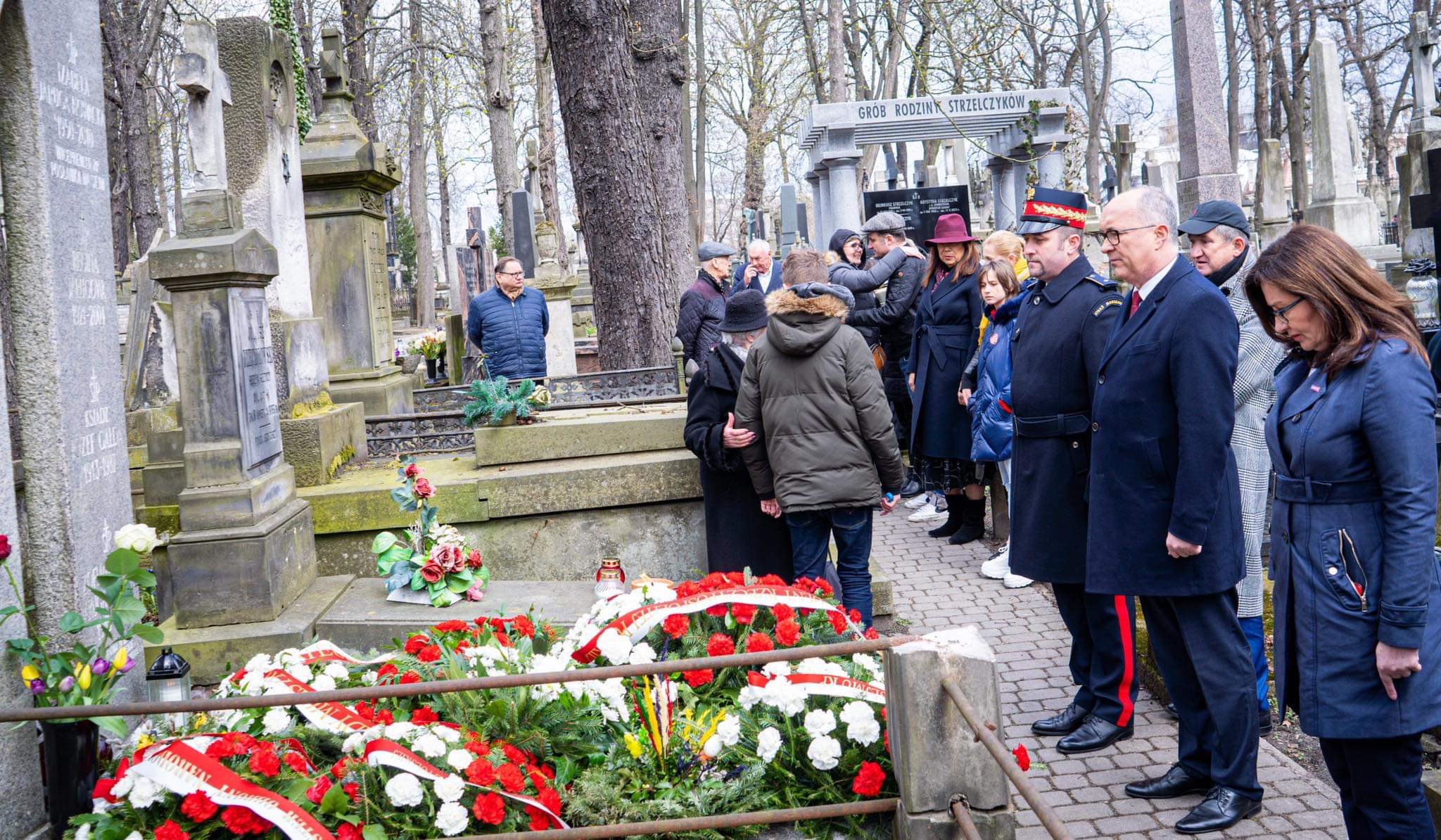 Grupa osób na cmentarzu składa kwiaty i oddaje hołd przy grobach podczas obchodów 12. rocznicy katastrofy smoleńskiej.
