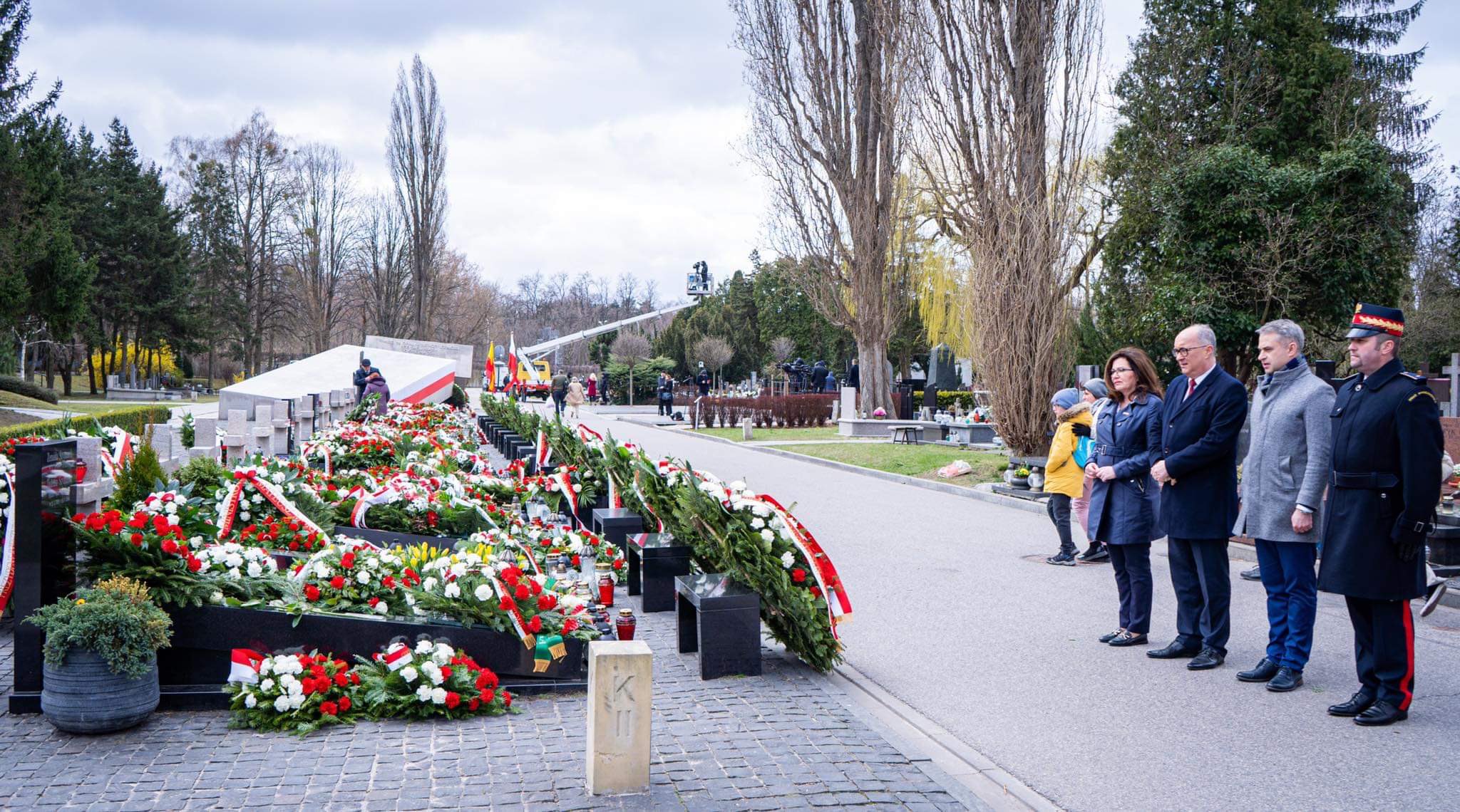 Grupa osób stojąca na cmentarzu podczas 12. rocznicy tragedii smoleńskiej, otoczona licznymi wieńcami i kwiatami w biało‑czerwonych barwach.