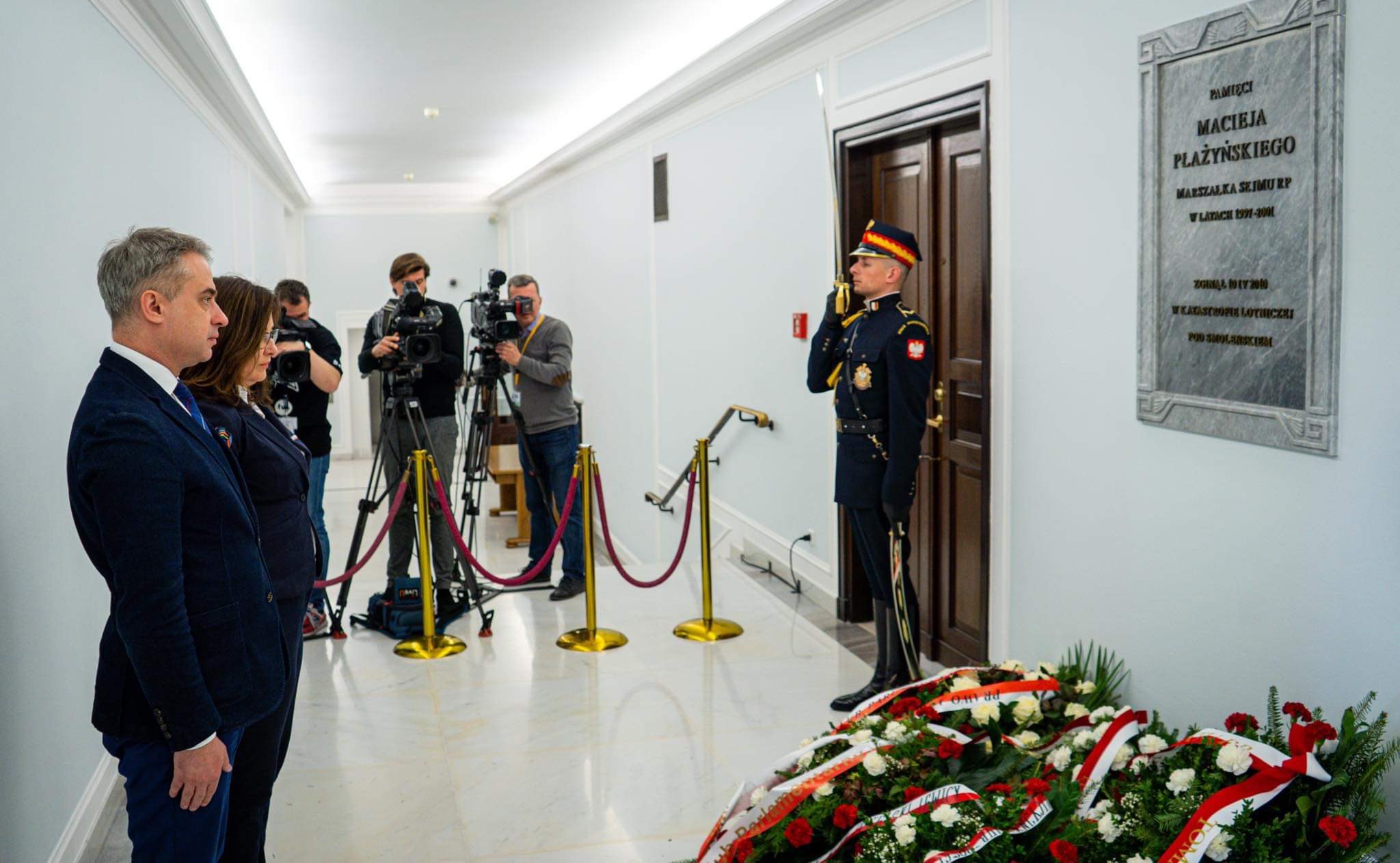 Korytarz, żołnierz w mundurze salutuje przy tablicy upamiętniającej ofiary tragedii smoleńskiej; obok leżą wieńce, a grupa dziennikarzy z aparatami fotografuje wydarzenie.