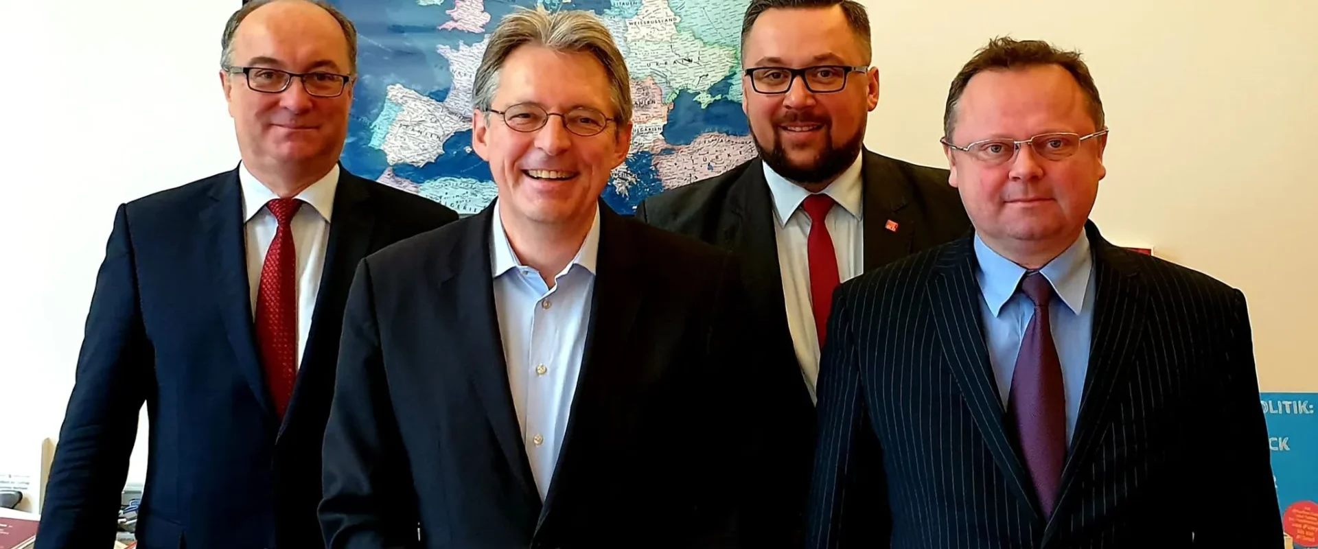 Czterech mężczyzn w garniturach stoi w biurze z dużą mapą świata w tle, podczas spotkania delegacji SLD ze SPD w Berlinie na temat współpracy i wyborów do europarlamentu.