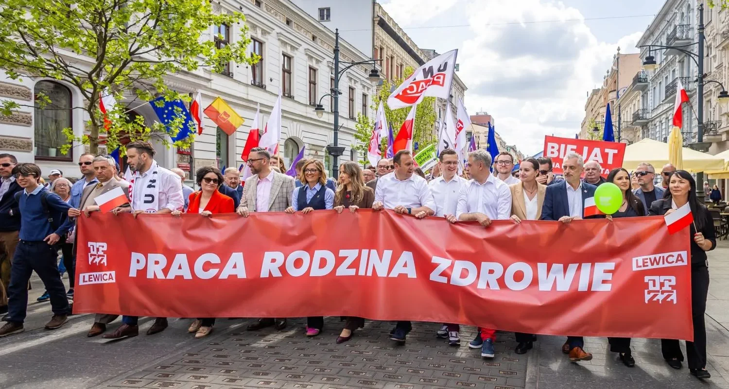 Grupa ludzi maszeruje ulicą podczas parady z czerwonym banerem z napisem PRACA RODZINA ZDROWIE i flagami, w kontekście Święta Pracy 2025.