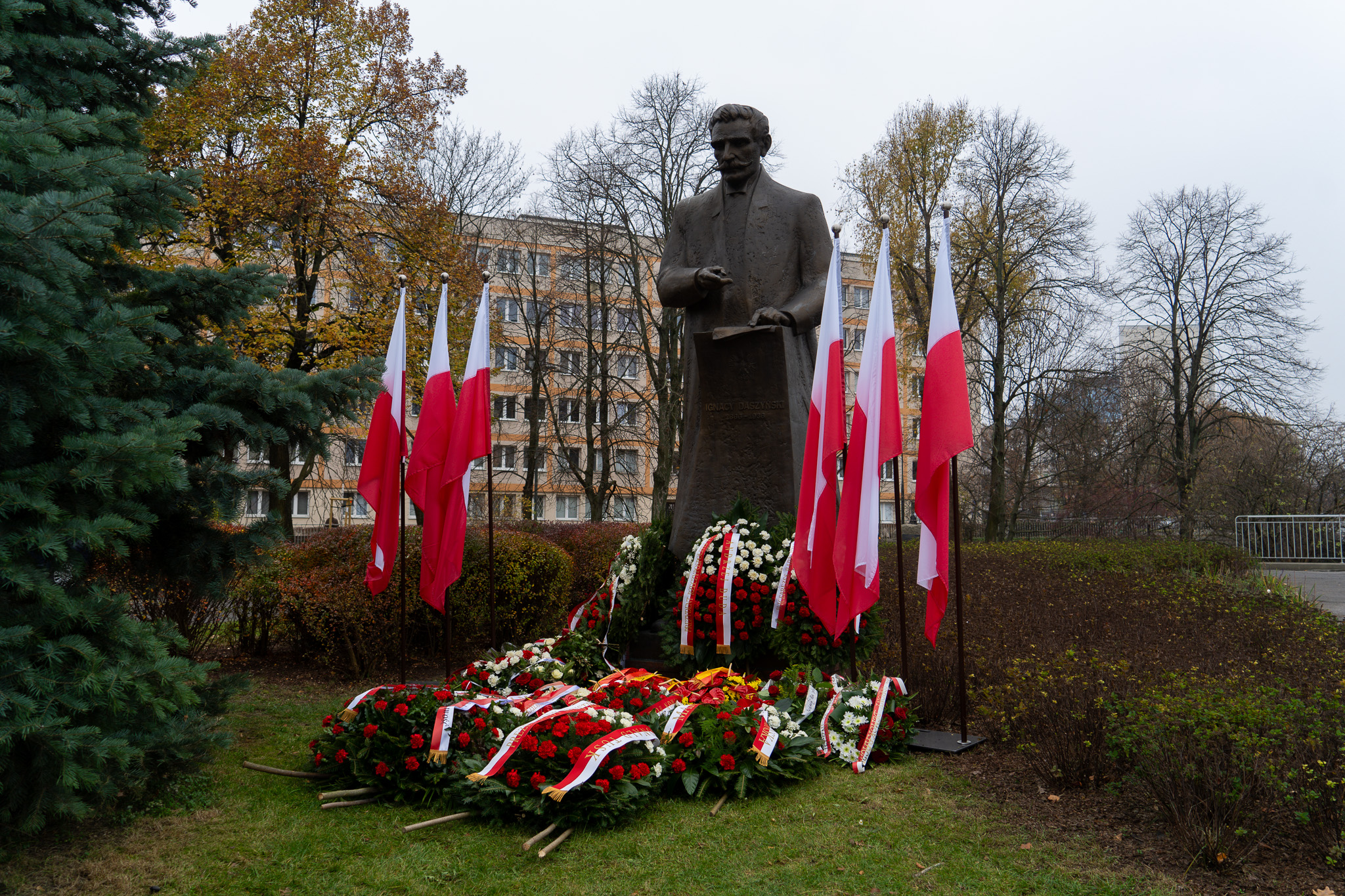 Posąg mężczyzny w parku otoczony flagami Polski i wieńcami z czerwonych i białych kwiatów, upamiętniający Narodowe Święto Niepodległości.