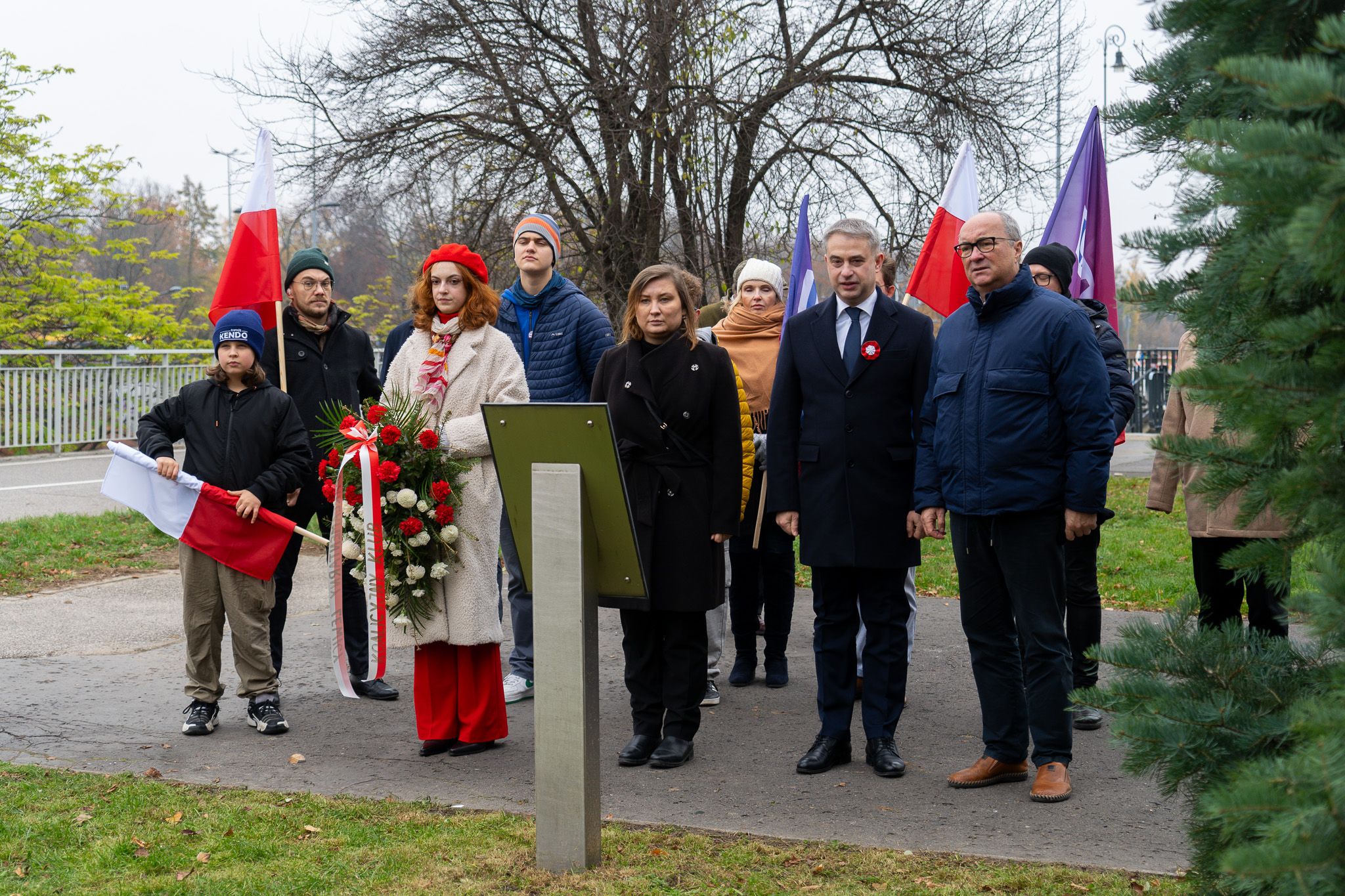 Grupa ludzi podczas obchodów Narodowego Święta Niepodległości stoi na alei z flagami Polski, obok wieńca z czerwonych i białych kwiatów przy tablicy pamiątkowej.