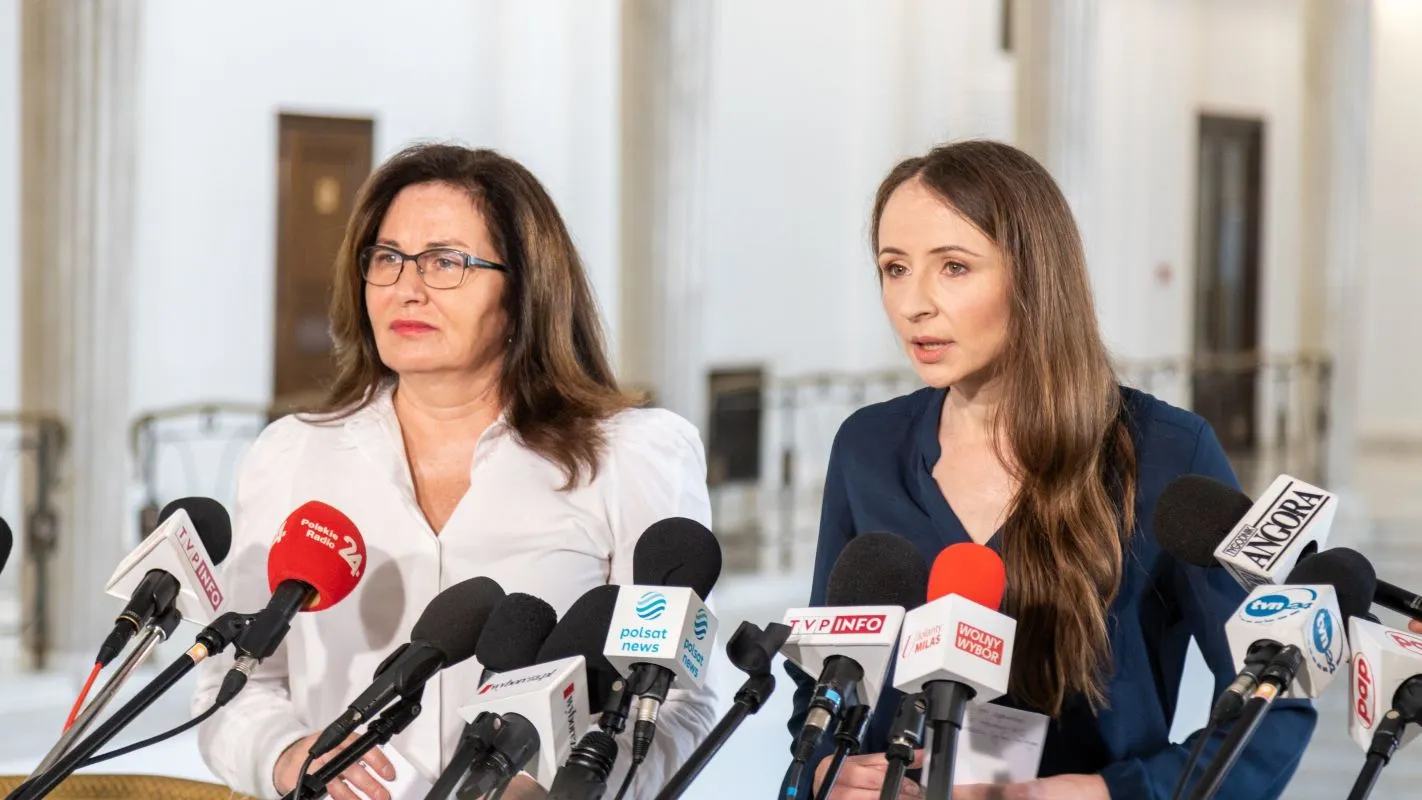 Na zdjęciu widoczne są dwie kobiety stojące przed mikrofonami podczas konferencji prasowej.