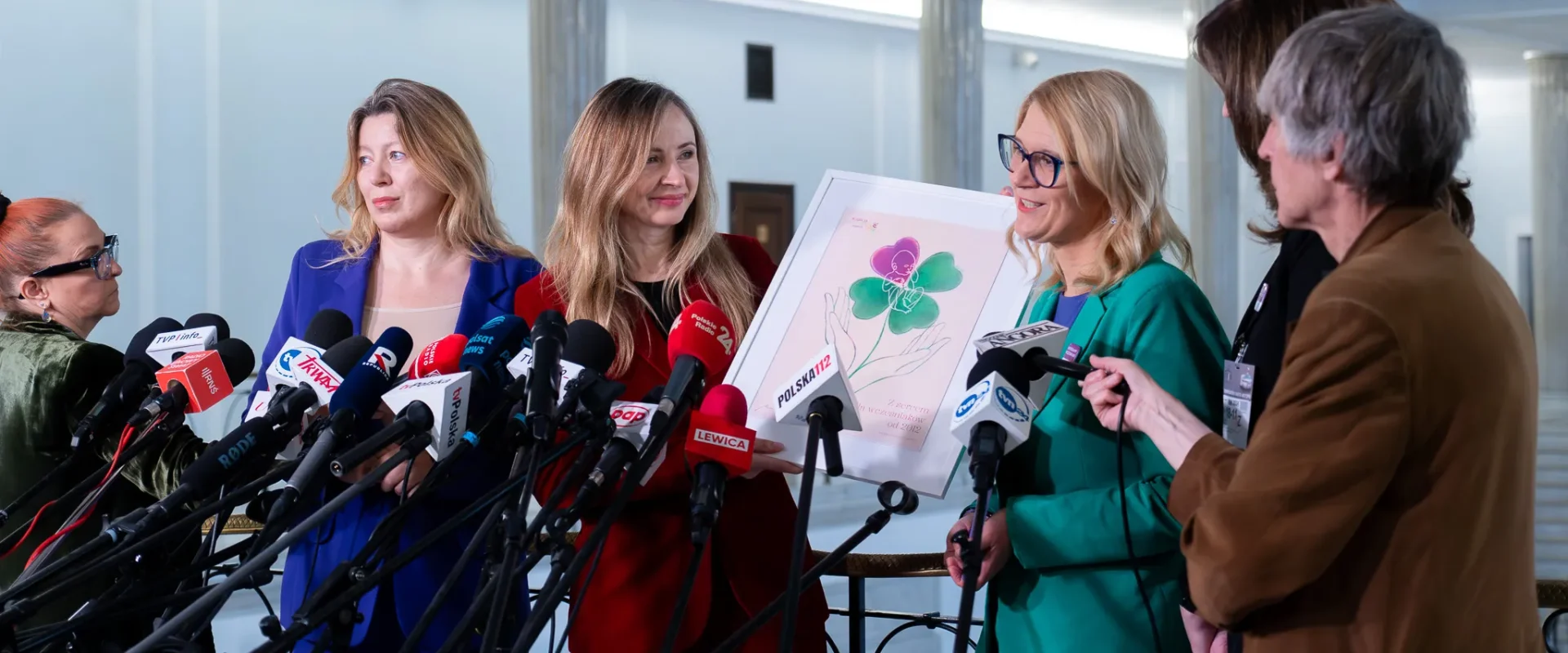 Grupa kobiet na konferencji prasowej stoi obok licznych mikrofonów, prezentując ramkę z ilustracją dotyczącą dłuższego urlopu macierzyńskiego dla rodziców wcześniaków.