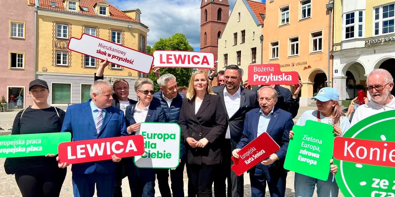 Grupa ludzi stojąca na miejskim placu w kolorowych kamienicach trzyma banery i hasła protestujące przeciwko produkcji biomasy z drzew.