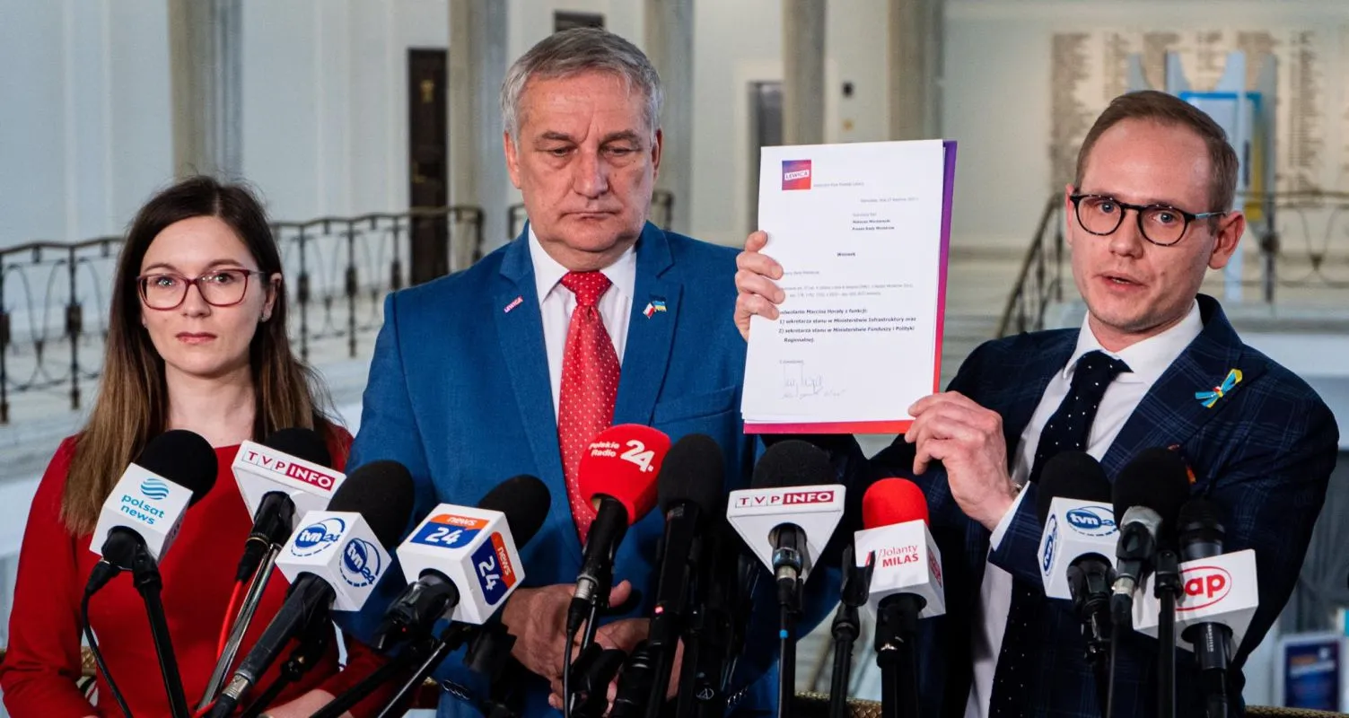 Trzy osoby na konferencji prasowej; mężczyzna w niebieskim garniturze z czerwonym krawatem trzyma dokument, obok niego drugi mężczyzna w okularach go prezentuje, a po lewej stoi kobieta w czerwonej bluzce, przed licznymi mikrofonami.