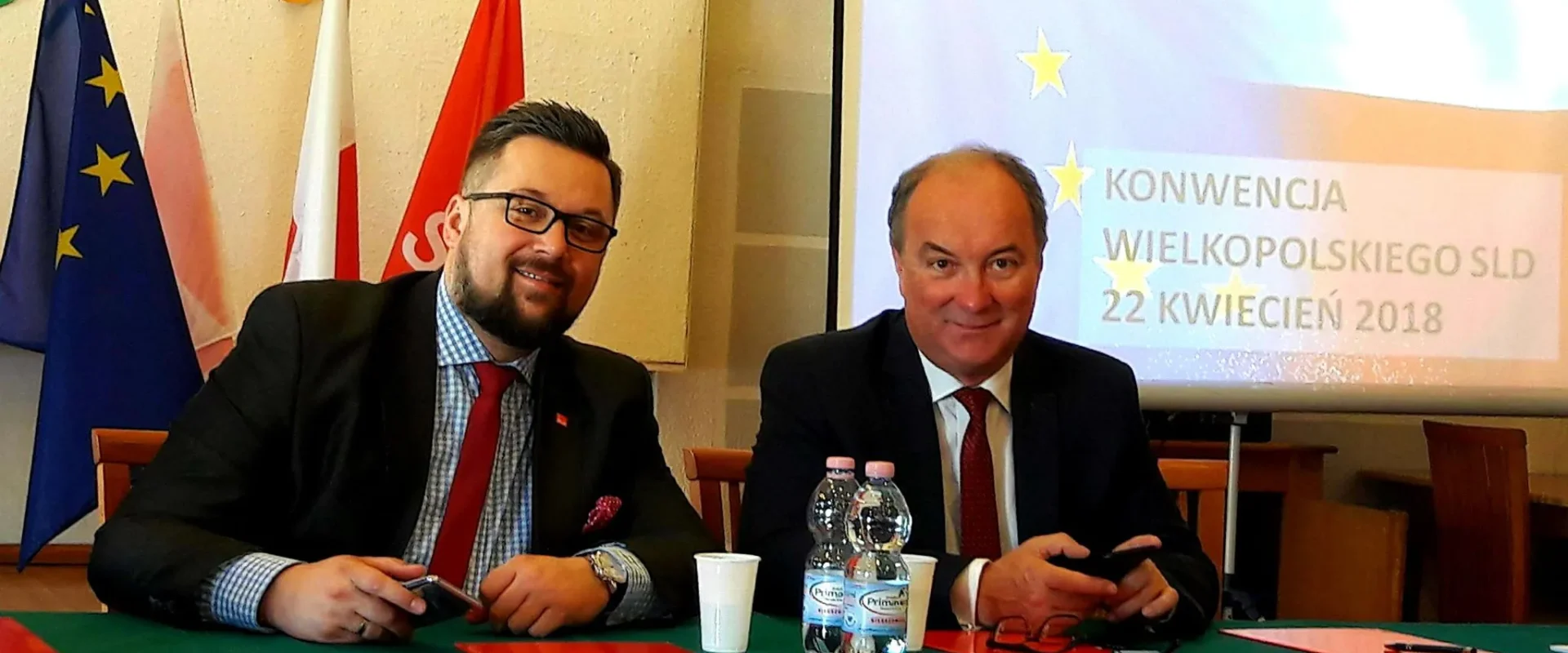 Dwóch mężczyzn w garniturach siedzi przy zielonym stole podczas konferencji, z flagami w tle i projekcją w którą widać napis Konwencja Wielkopolskiego SLD 22 kwietnia 2018.